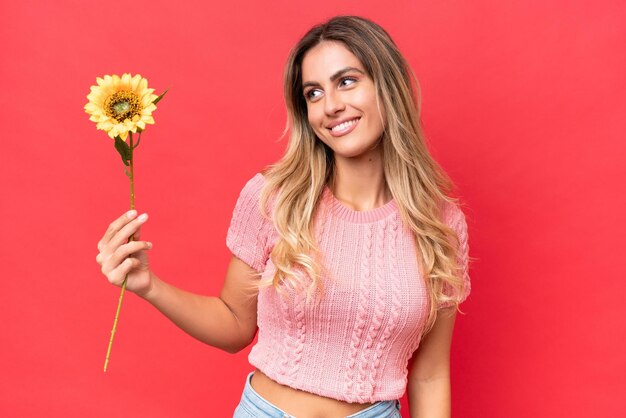 Junge hübsche uruguayische Frau, die Sonnenblume isoliert auf dem Hintergrund mit glücklichem Ausdruck hält