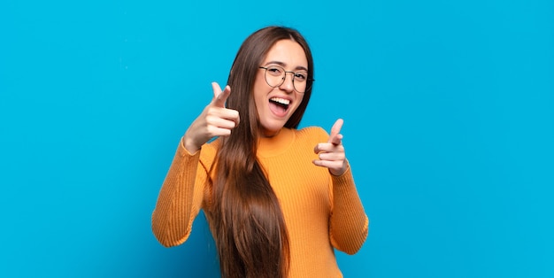 Junge hübsche zufällige Frau, die mit einer positiven, erfolgreichen, glücklichen Haltung lächelt und zeigt, Waffenzeichen mit den Händen