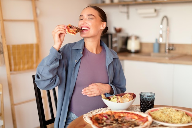 Junge hungrige schwangere Frau beißt ein Stück Pizza und hat Lust auf Junkfood, die am vollen Tisch sitzt