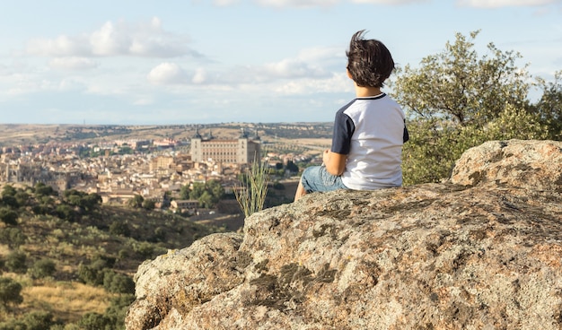 Junge im blauen und weißen Hemd, das auf einem großen Stein mit seinem Rücken sitzt und die Landschaft betrachtet