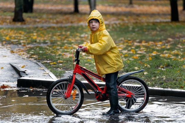 Junge im gelben Regenmantel auf dem Fahrrad fährt auf Pfützen. Regnerischer Herbsttag.