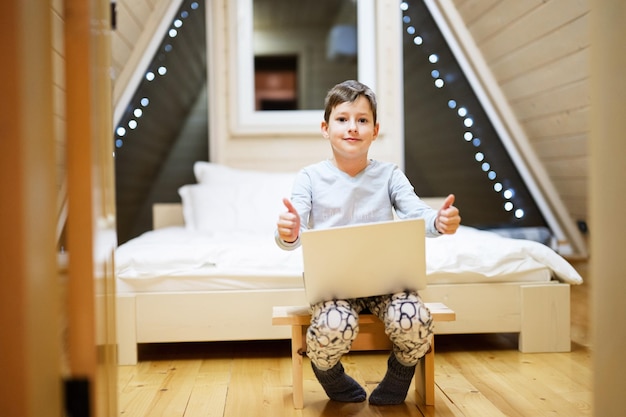 Junge im Pyjama sitzen mit Laptop in der Holzhütte zu Hause Konzept des Glücks der Freizeitbeschäftigung in der Kindheit