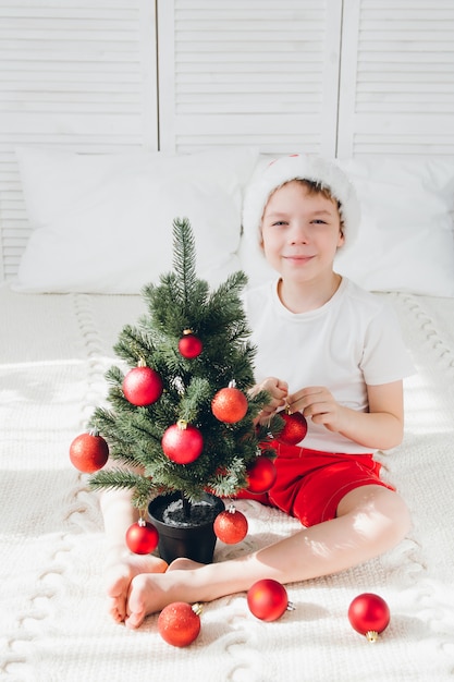 Junge im roten Sankt-Hut verziert kleine Christbaumkugeln