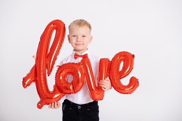 Junge in Anzug und roter Fliege hält die Inschriftenliebe von Luftballons an einer weißen Wand.