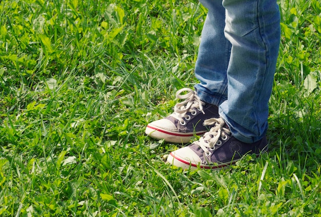 Junge in blauen Turnschuhen und Jeans, die im Sommer auf dem sonnigen Tag des grünen Grases stehen Nahaufnahme des getönten Bildes des Beines