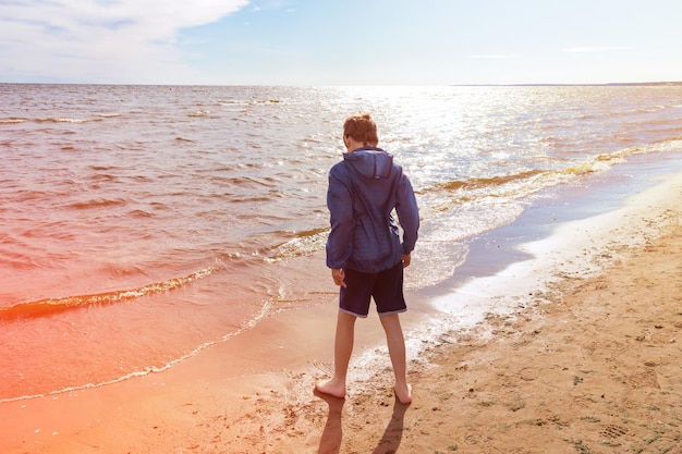 Junge in blauer Jacke und Shorts barfuß auf Sand am Meer, sonniger Tag