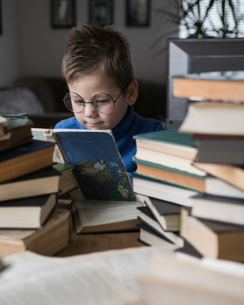 Junge in Brille liest ein Buch mit einem Stapel Bücher neben sich.