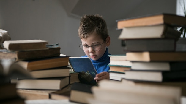 Junge in Brille liest ein Buch mit einem Stapel Bücher neben sich.