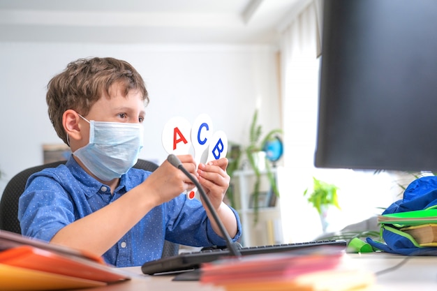 Junge in der Gesichtsmaske unter Verwendung des Computers, Hausaufgaben während der Coronavirus-Quarantäne machend