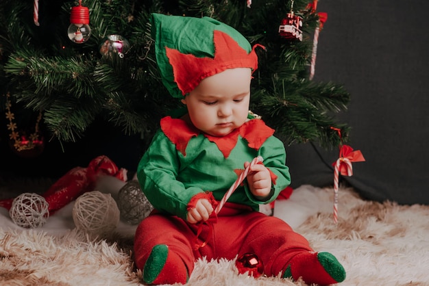 Junge in einem Elfenkostüm sitzt unter einem Weihnachtsbaum mit einem Lutscher