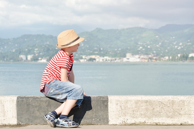 Junge in einem Hut und in einem gestreiften T-Shirt, die auf dem Strand sitzen und das Meer betrachten. Seitenansicht
