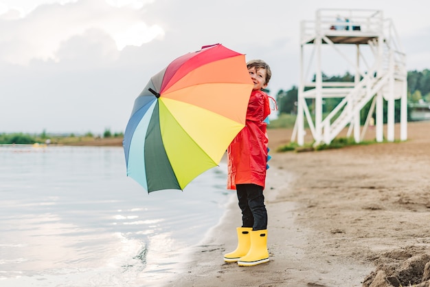 Junge in einem roten Regenmantel und gelben Gummistiefeln steht am Flussufer und hält einen Regenbogenregenschirm