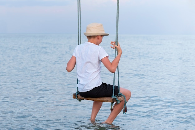 Junge in einem Strohhut, der auf Seil sitzt, schwingt über Wasser