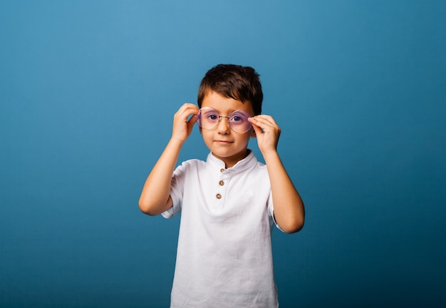 Junge in einem weißen T-Shirt hält eine Brille in der Hand und schaut auf blauem Hintergrund in die Kamera. Junge mit Brille.