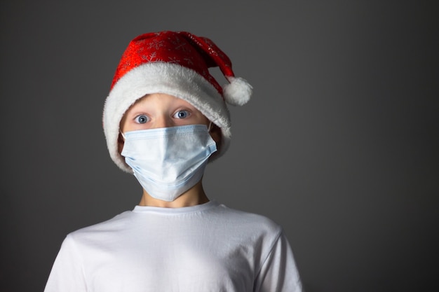 Junge in einem weißen T-Shirt, Weihnachtsmütze und medizinische Maske auf grauem Hintergrund.