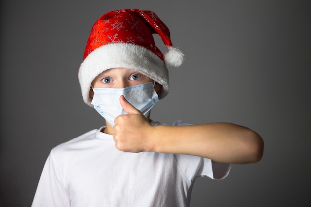 Junge in einem weißen T-Shirt, Weihnachtsmütze und medizinischer Maske zeigt einen Daumen auf Grau