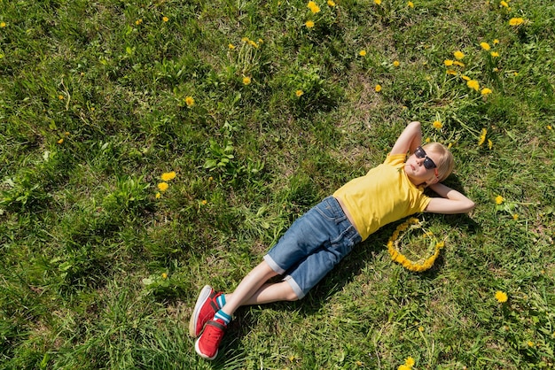 Junge in gelbem T-Shirt und Jeans-Shorts mit den Händen hinter dem Kopf liegt auf dem Gras und ruht sich auf.