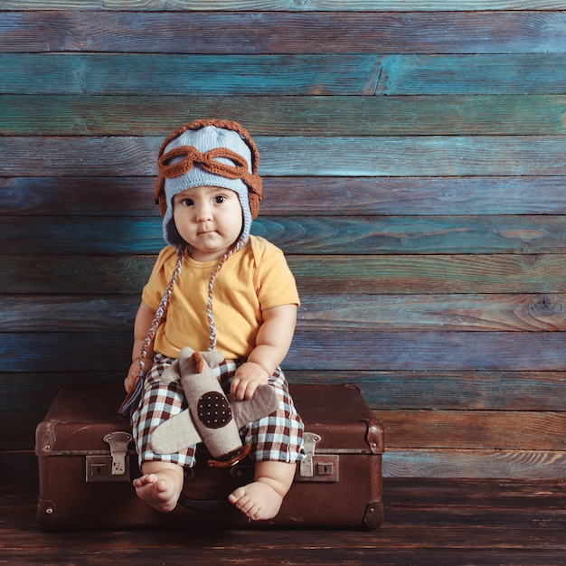 Junge in gestrickter Pilotenmütze spielt mit Spielzeugflugzeug, sitzt auf einem altmodischen Koffer