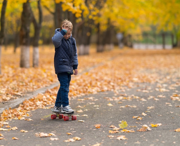 Junge in grauer Jacke Skateboarding im Park in der Herbstsaison, von vorne gesehen. Kopieren Sie den Raum mit verschwommenen Bäumen und gelben Blättern zur Seite.