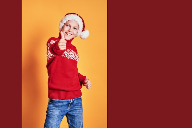 Junge in Sankt-Hut und in roter Strickjacke, die Spaß haben und sich Daumen am Weihnachtstag gegen gelben Hintergrund zeigen