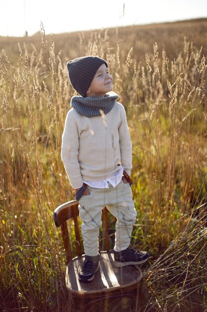 Junge in warmer Kleidung steht auf einem Stuhl auf einem Weg auf einem Feld mit getrocknetem Gras im Herbst