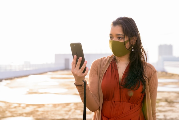 Junge Inderin mit Maske unter Verwendung des Telefons am Dach des Gebäudes