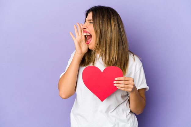 Junge indische Frau, die ein Herz des Valentinstags lokalisiert schreit und Palme nahe geöffnetem Mund hält.