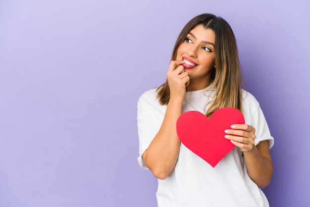 Junge indische Frau, die ein Valentinstagherz hält, lokalisiert entspanntes Denken an etwas, das einen Kopienraum betrachtet.