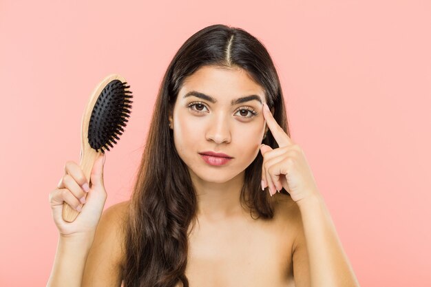 Junge indische Frau, die eine Haarbürste hält, die seine Schläfe mit dem Finger zeigt, denkend, konzentrierte sich auf eine Aufgabe.