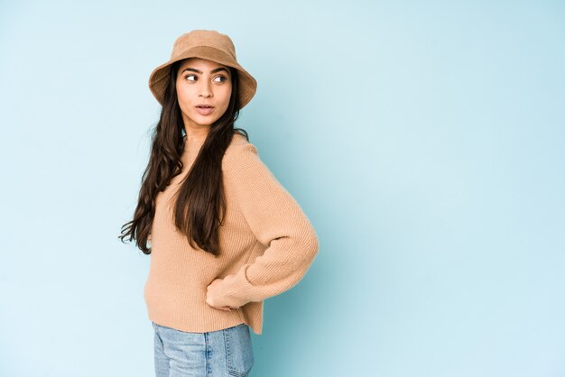 Junge indische Frau, die einen Hut auf blauer Wand trägt, schaut lächelnd, fröhlich und angenehm zur Seite.