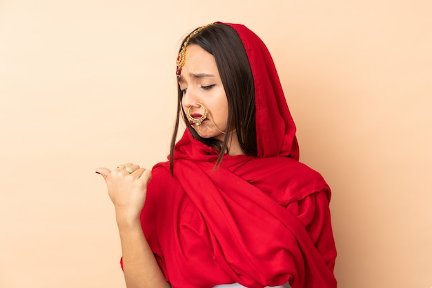 Junge indische Frau lokalisiert auf beige Wand unglücklich und zeigt zur Seite