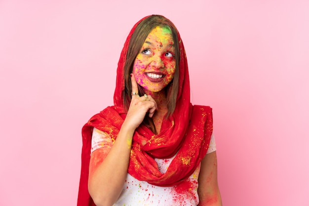 Junge indische Frau mit bunten Holi-Pulvern auf ihrem Gesicht auf rosa Wand, die eine Idee beim Nachschlagen denkt