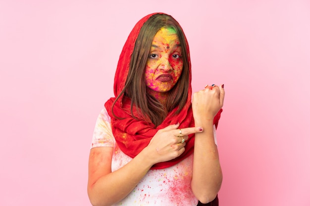 Junge indische Frau mit bunten Holi-Pulvern auf ihrem Gesicht lokalisiert auf rosa Wand, die die Geste der Verspätung macht