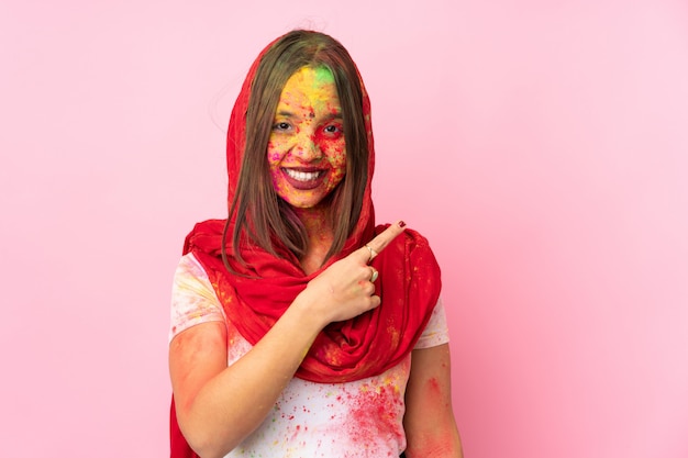 Junge indische Frau mit bunten Holipulvern auf ihrem Gesicht auf rosa Wand, die zur Seite zeigt, um ein Produkt zu präsentieren