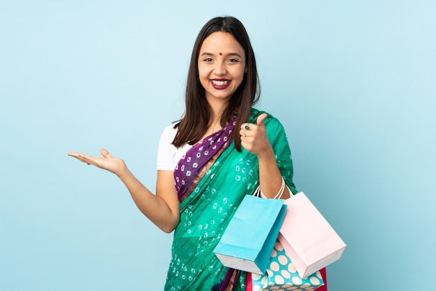Junge indische Frau mit Einkaufstüten, die imaginären Copyspace auf der Handfläche halten, um eine Anzeige und mit den Daumen nach oben einzufügen