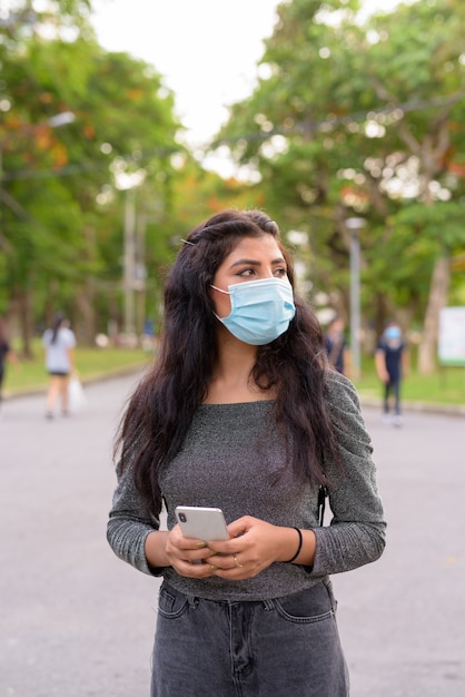 Junge indische Frau mit Maskendenken beim Telefonieren im Park im Freien
