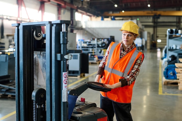 Junge Ingenieurin, die einen elektrischen Gabelstapler fährt, während sie sich entlang des Lagers bewegt