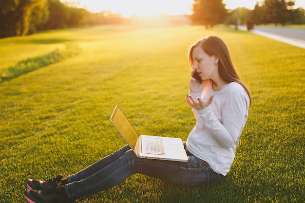 Junge intelligente Studentin in Freizeitkleidung. Frau, die am Handy spricht, an Laptop-PC-Computer im Stadtpark auf grünem Grassonnenscheinrasen im Freien arbeitet. Mobiles Büro. Freiberufliches Geschäftskonzept