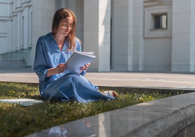 Junge intelligente Studentin sitzt auf Gras in der Nähe des Universitätsgebäudes und studiert und liest mit Büchern und...