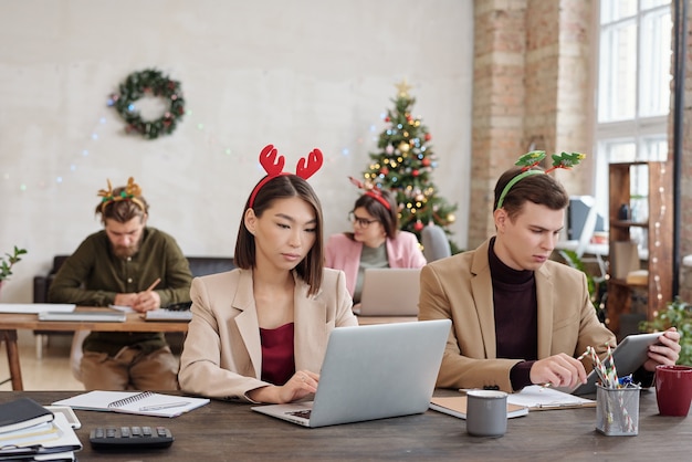 Junge interkulturelle Mitarbeiter in Smart Casualwear und Weihnachtsstirnbändern vernetzen sich vor Laptops in Reihen sitzend