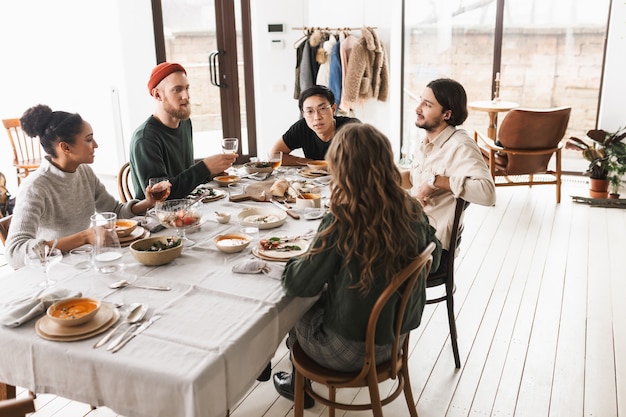 Junge internationale Freunde sitzen am Tisch voller Essen und unterhalten sich träumerisch