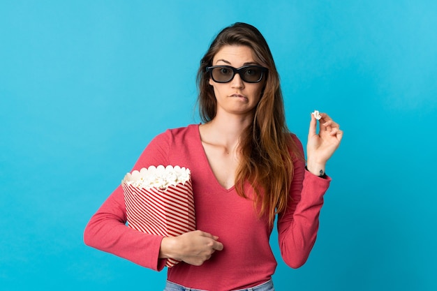 Junge Irlandfrau isoliert auf blauem Hintergrund mit 3D-Brille und hält einen großen Eimer Popcorn