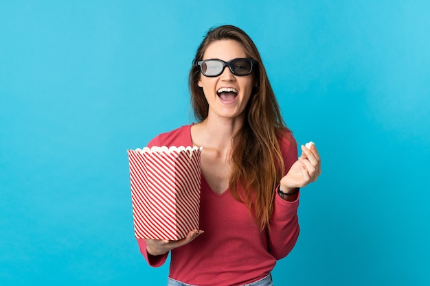 Junge Irlandfrau lokalisiert auf blauem Hintergrund mit 3D-Brille und hält einen großen Eimer Popcorn
