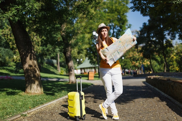 Junge irritierte Reisende Touristenfrau in gelber Sommerkleidung, Hut mit Kofferstadtplan gehen in der Stadt im Freien. Mädchen, das ins Ausland reist, um am Wochenende zu reisen. Tourismus Reise Lebensstil.