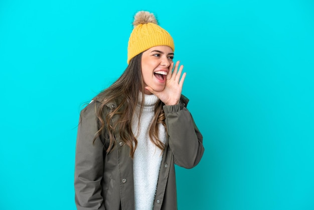 Junge Italienerin mit Winterjacke und Hut isoliert auf blauem Hintergrund schreiend mit weit geöffnetem Mund zur Seite