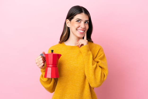 Junge italienische Frau, die eine Kaffeemaschine isoliert auf rosa Hintergrund hält und eine Idee denkt, während sie nach oben schaut