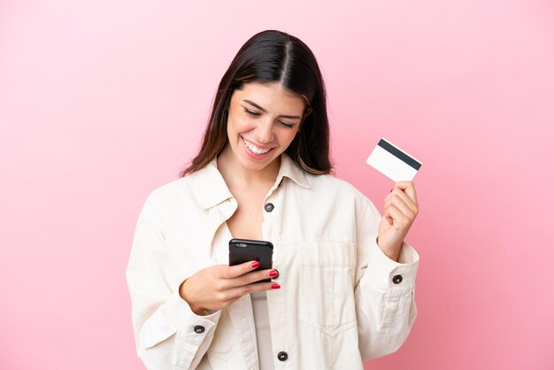 Junge italienische Frau isoliert auf rosafarbenem Hintergrund, die mit dem Handy mit einer Kreditkarte kauft