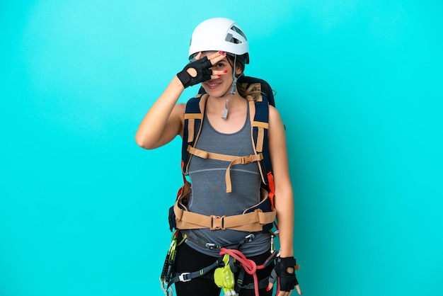 Junge italienische Kletterin isoliert auf blauem Hintergrund, die die Augen mit den Händen bedeckt und lächelt