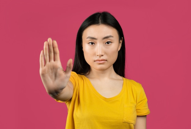 Junge japanische Dame, die STOP gestikuliert und eine Ablehnungsgeste zeigt, die ihre negative Einstellung auf rosafarbenem Hintergrund ausdrückt