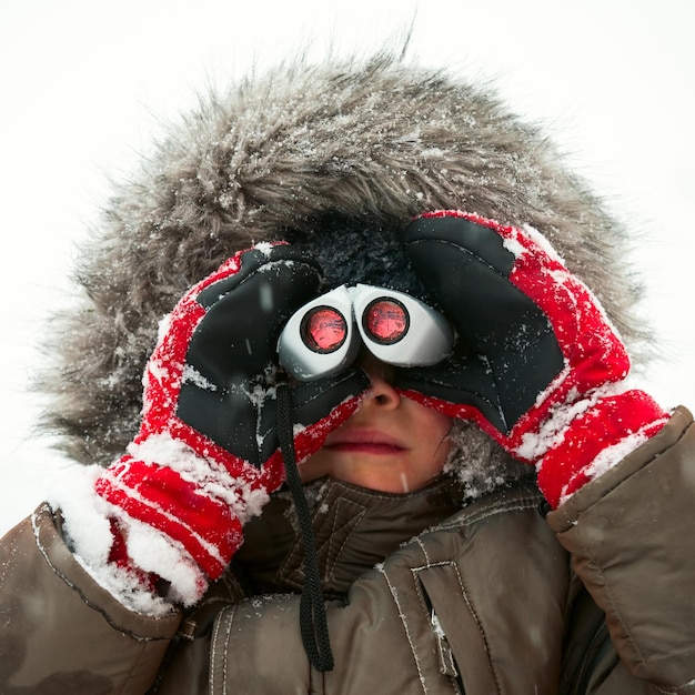 Junge Junge trägt eine Winterjacke mit pelziger Kapuze und roten Handschuhen und schaut durch ein Fernglas